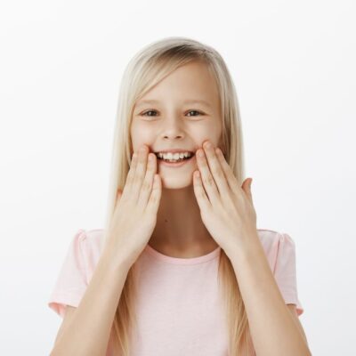 pleased grinning adorable child with blond hair smiling broadly holding palms near lips being amazed satisfied with healthy teeth attending dentist feeling happiness min درمانگاه کودک سالم